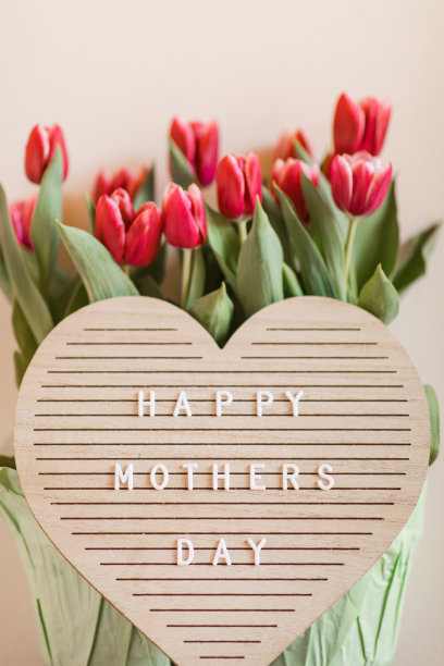 一个心形的字母板，上面写着“母亲节快乐”，明亮的粉红色郁金香盛开，郁郁葱葱的绿色茎和叶子在它后面，在明亮的自然日光下拍摄，中性的米色背景与复制空间图片下载