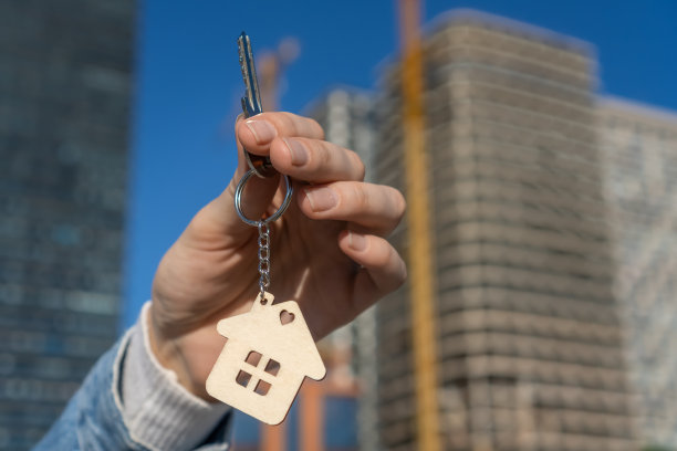 手拿小房子形状的钥匙链和钥匙。以典型的美国乡村风格的房子为背景。建造、出售或购买公寓。开发商、房地产经纪人或房地产中介。图片下载