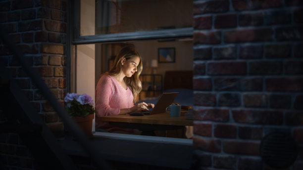 金发美女坐在卧室的办公桌前用笔记本电脑工作。女性职业自由职业者晚上在舒适的家里工作。窗外的景色图片下载