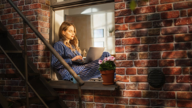 在家办公概念:年轻的女商人使用笔记本电脑，穿着睡衣在卧室的窗台上远程工作。女人喜欢在舒适的公寓里完成工作任务图片下载