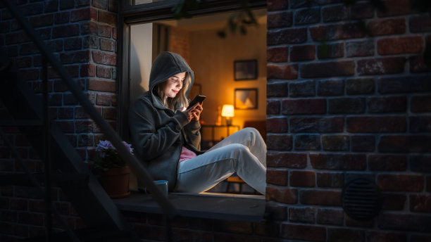 在黑暗中坐在窗边专心地用智能手机打字的女人。网瘾女性使用手机应用进行网上购物、社交媒体发帖、上网浏览图片下载