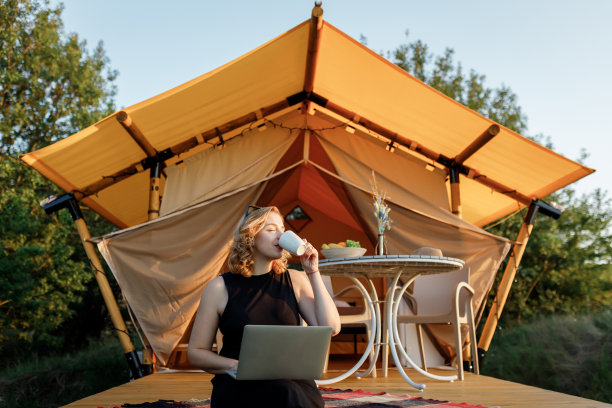 快乐的女人自由职业者使用笔记本电脑和喝咖啡在一个舒适的豪华帐篷在一个阳光明媚的日子。豪华户外露营帐篷，适合夏季度假度假。生活方式的概念图片下载