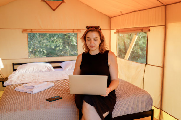 在一个阳光明媚的日子里，在舒适的豪华露营帐篷里使用笔记本电脑的微笑女性自由职业者。豪华户外露营帐篷，适合夏季度假度假。生活方式的概念图片下载