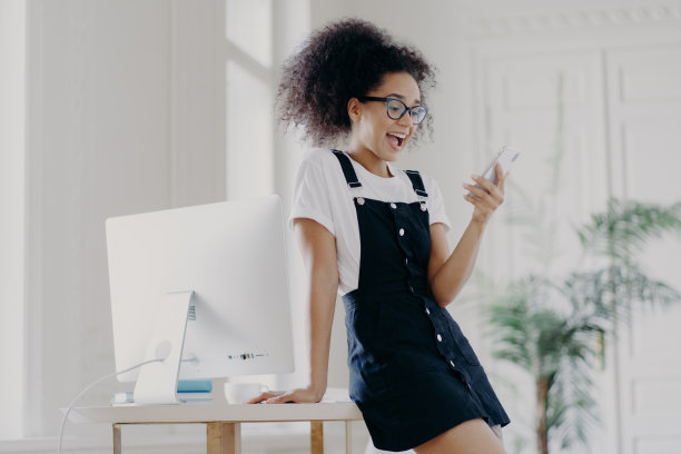 室内照片，惊喜的年轻非洲裔美国妇女戴着眼镜，白色t恤和沙拉芬，用手机上网聊天，在电脑旁摆姿势。图片下载