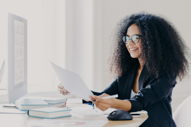 快乐的非洲裔美国妇女，卷发，做财务报告，拿着纸质文件，有快乐的微笑，戴着矫正视力的眼镜，穿着优雅的衣服，电脑屏幕在前面图片下载