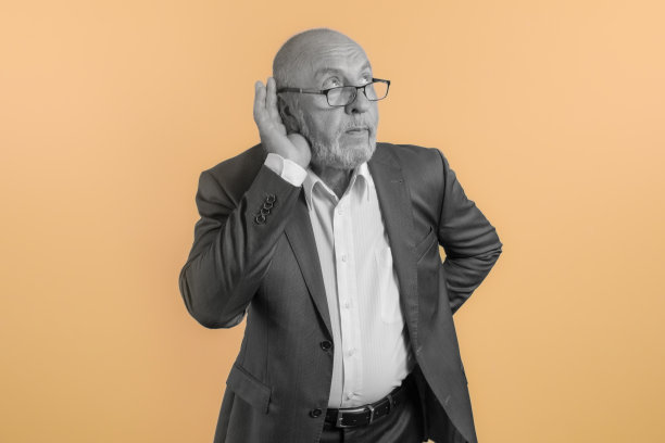 一个老人摆出偷听谈话的姿势。一个试图倾听的人……手放在耳朵上。明亮的橙色背景。艺术图片下载