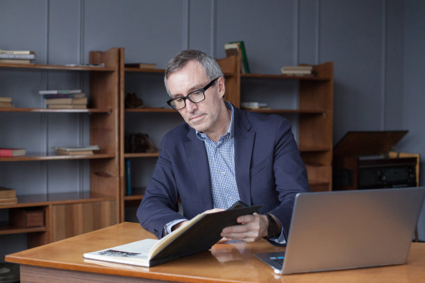 一个穿着西装的成熟商人的肖像，坐在笔记本电脑前，在办公室里沉思地读着书图片下载