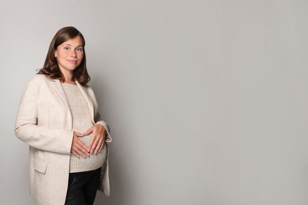 漂亮的年轻孕妇站在灰色的工作室墙壁背景下，用手在肚子上做心脏图片下载