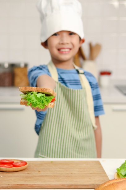 手头的三明治特写，带着围裙和厨师帽的快乐微笑的亚洲男孩作为模糊的背景，孩子在厨房准备简单的饭菜，孩子厨师喜欢自己做饭和吃。图片下载