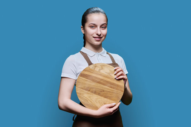 年轻的女学生服务员系着围裙，蓝色背景上有一个空的木制托盘图片下载