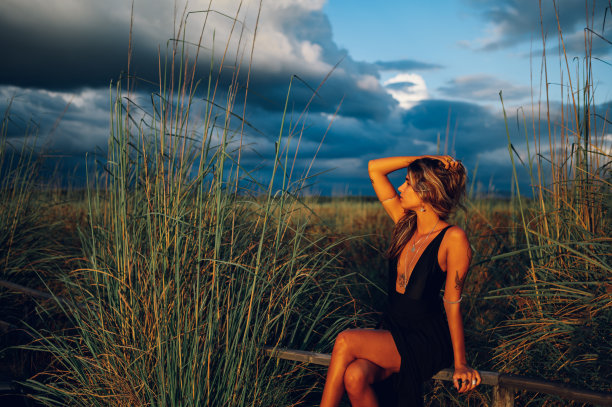 一位皮肤黝黑、身上有纹身的年轻女子穿着优雅的夏装，坐在大自然的篱笆上。背景是暴风雨，戏剧性的天空。图片下载