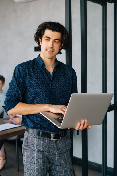 身着休闲服、深思熟虑的商人的垂直肖像，手里拿着笔记本电脑，站在办公室里，望向别处。创业团队讨论项目图片下载