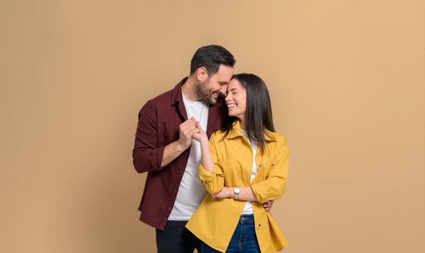 这是一对深情微笑的年轻夫妇的肖像，他们面对面地手牵着手，站在米色的背景上。相爱的男人和女人闭着眼睛浪漫地摆姿势图片下载