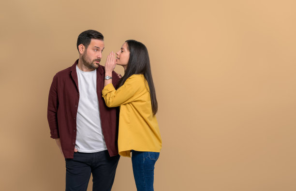 严肃的男友独自站在米色的背景上，看着女友在耳边窃窃私语。年轻男女互相说闲话图片下载