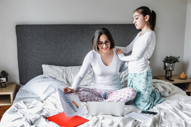 拉丁美洲:母亲躺在床上用笔记本电脑工作，女儿躺在床上在家办公图片下载