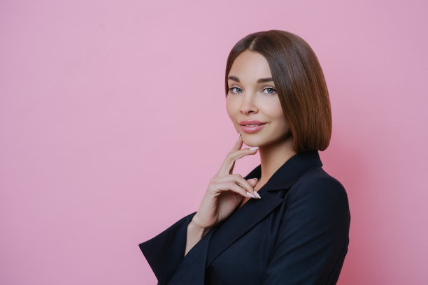 工作室拍摄的迷人的年轻女企业家穿着正装，抚摸着下巴，皮肤健康，化着妆，侧身站在镜头前，孤立在玫瑰色的工作室背景下图片下载