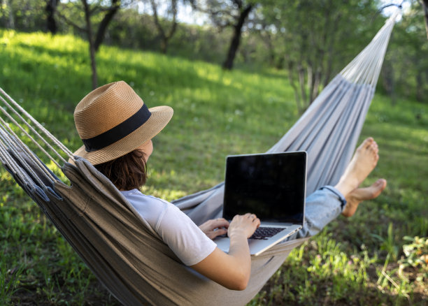 一位戴着帽子的年轻女子躺在夏日花园的吊床上使用笔记本电脑。夏天的生活方式图片下载