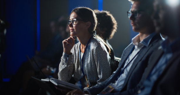 一位热情的年轻女性参加了一场励志演讲活动，她坐在拥挤的音乐厅里，靠向舞台，在笔记本上写下励志名言。图片下载