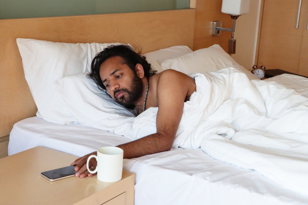 印度男子从羽绒被下穿过酒店的双人床，拿着咖啡杯关掉床头柜上的智能手机闹钟，重点放在前景上图片下载