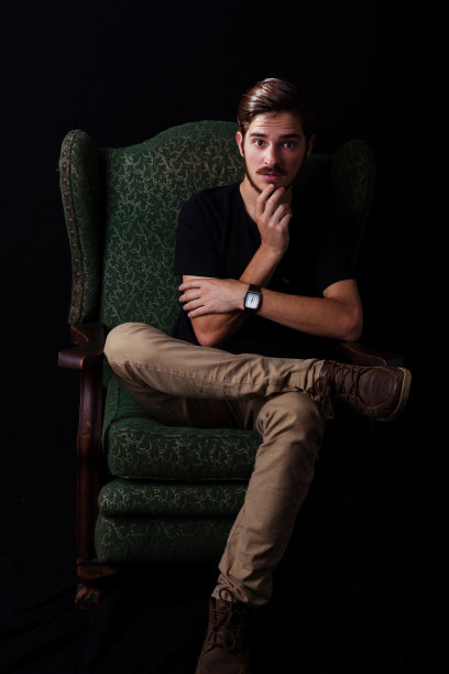 年轻的成年男子坐在老式的靠背椅子上，手放在下巴上，表情险恶或冲突。单调，黑白为戏剧效果。表演学校的面部表情概念。图片下载