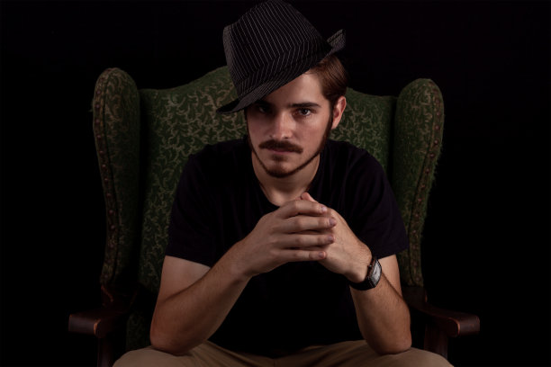 年轻成年男性的近照，看起来阴险或沉思。颜色暗色调戏剧性的效果，黑暗和喜怒无常的系列。概念形象的魔术师在翼背椅不高兴和诡计。图片下载