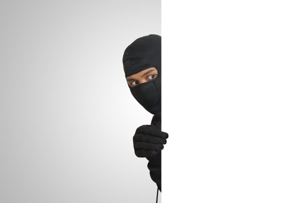 神秘的小偷穿着黑色连帽衫躲在墙后，偷偷摸摸，寻找被盗物品。犯罪的概念。灰色背景上的孤立图像图片下载