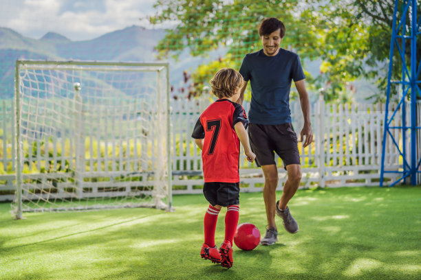 可爱的小男孩穿着红色的足球服和他的教练或父亲踢足球，在球场上，在户外。活跃的孩子与孩子或父亲一起运动，微笑的快乐男孩在夏天玩得开心图片下载