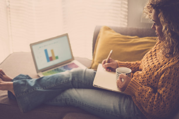 一位在家工作的妇女躺在沙发上舒适地写着笔记，同时使用无线连接的笔记本电脑。现代女性与新常态的远程办公智能办公商务图片下载