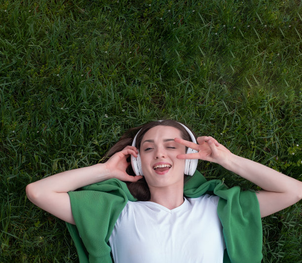 一个十几岁的女孩戴着耳机听音乐，躺在公园的绿草地上，微笑着露出舌头，穿着白色t恤和绿色毛衣，夏天的心情是一种生活方式图片下载