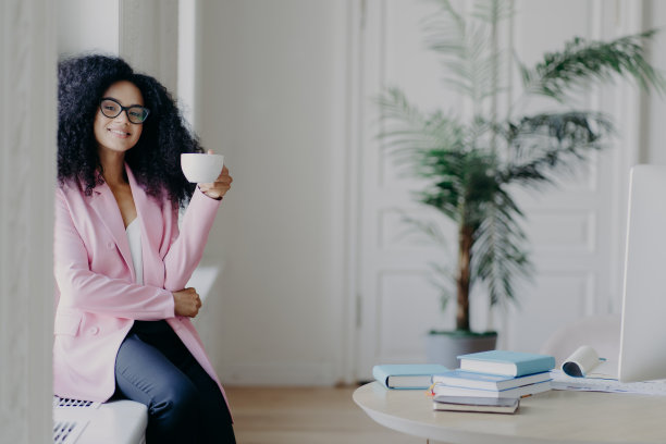积极的非裔美国女商务工作者坐在窗台前，端着咖啡杯，穿着正装，摆着摆着书的桌面，背景是绿色的室内植物，休息一下图片下载