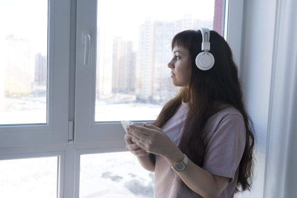 年轻女子戴着无线耳机，用手机听音乐，看着窗外。图片下载