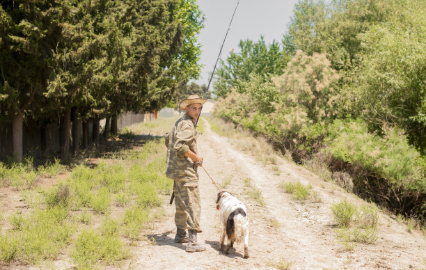 在阳光明媚的日子里，穿着迷彩服的沉思的男渔夫带着忠实的狗走在泥泞的路上。图片下载