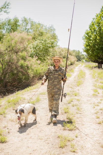 周末戴草帽、穿迷彩服的渔夫带着狗去钓鱼。图片下载