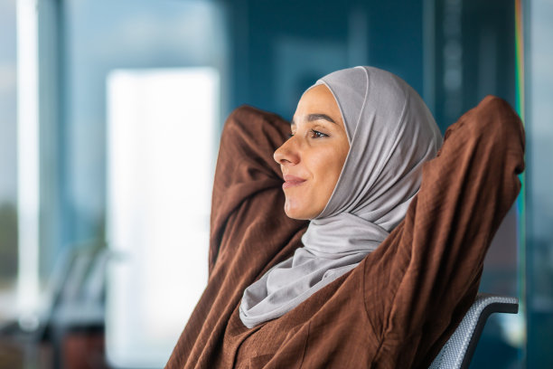 项目成功完成，工作场景：一位戴着头巾的年轻阿拉伯妇女在工作场所休息，若有所思地望着窗外图片下载