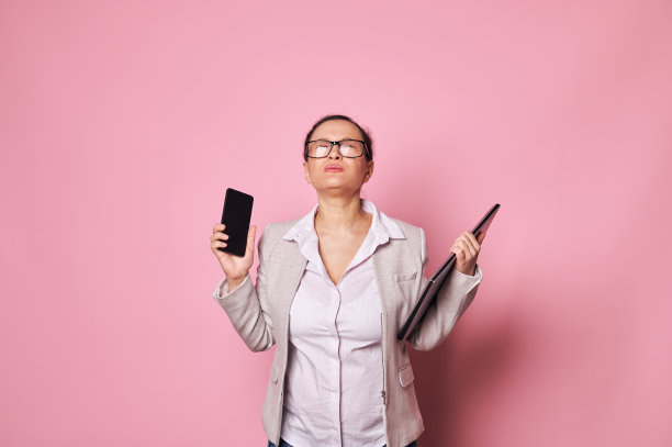 劳累过度、压力重重的怀孕女商人站在那里，粉红色的背景是笔记本电脑和手机。怀孕与事业图片下载