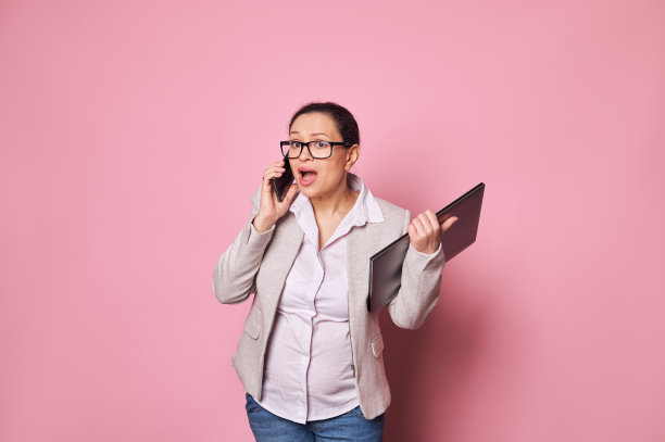 多任务处理怀孕的女商人被隔离在粉红色的背景中，拿着笔记本电脑用手机通话。图片下载