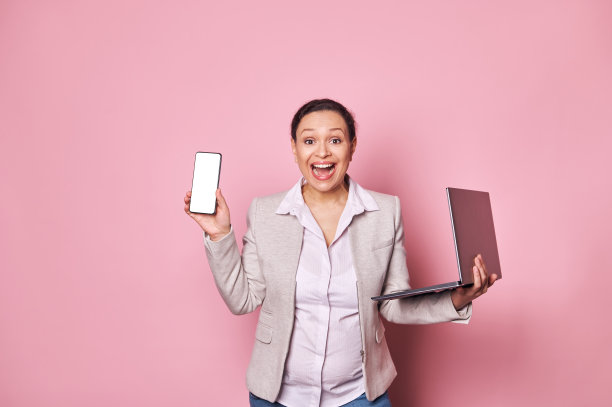 兴奋的怀孕商务女士表示惊讶和喜悦，摆着笔记本电脑和手机，白色屏幕的移动应用程序图片下载