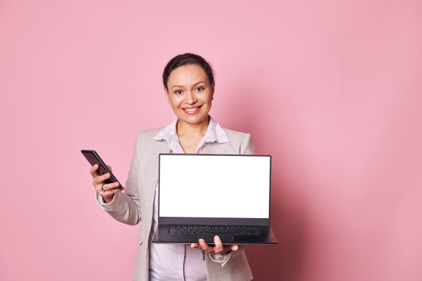 多民族积极的女人拿着智能手机，微笑着看着相机，同时展示着带有白色数字屏幕的笔记本电脑图片下载