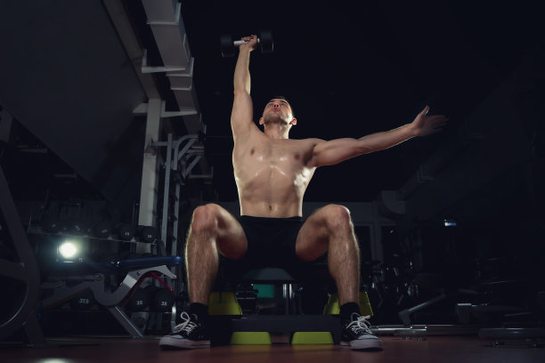 举起哑铃。肌肉发达的帅哥。运动和训练的概念。肌肉男在黑暗的健身房拿着哑铃，运动生活方式的概念。图片下载