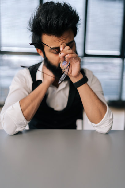 疲惫的印度商人按摩鼻梁，摘下眼镜，感觉眼睛疲劳，坐在办公桌前的垂直肖像。图片下载