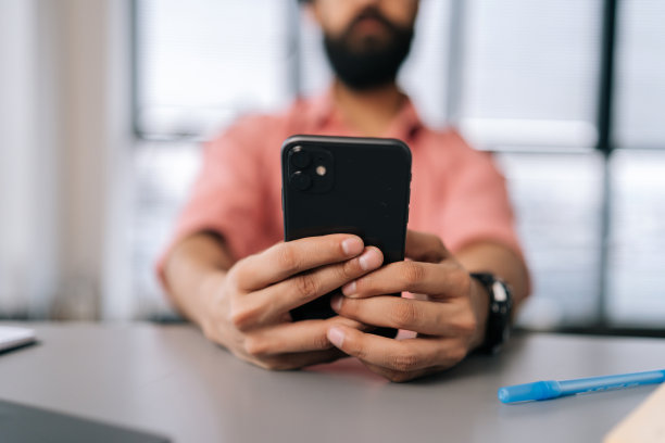 特写镜头，一个无法辨认的大胡子男人用智能手机坐在办公桌前，把书和工作文件放在窗前。图片下载