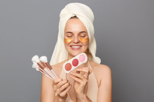 可爱可爱的美丽女人，眼睛下面有补丁，用毛巾裹着粉和刷子，孤立在灰色的背景下，欣赏她的新化妆美容产品。图片下载
