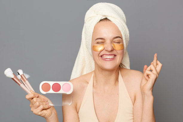 希望的女人眼睛下面有补丁，用毛巾裹着粉和刷子，交叉手指许愿，闭着眼睛，孤立在灰色的背景下。图片下载