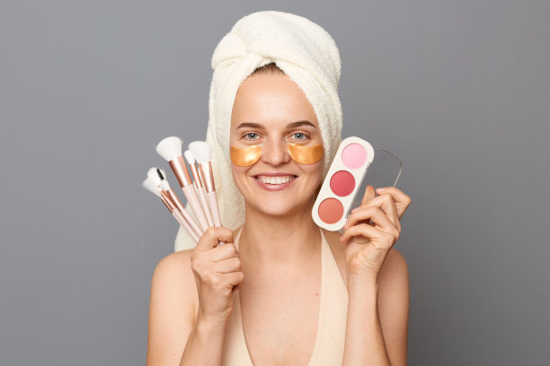 早上化妆。快乐快乐的女人，眼睛下面有补丁，用毛巾裹着，用刷子涂抹粉末，准备在灰色的背景上约会。图片下载