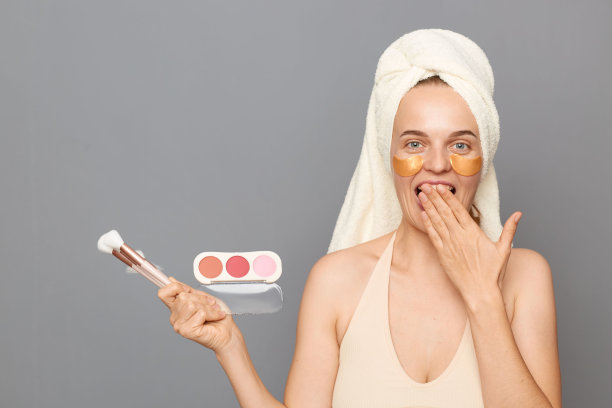 美容产品的惊人折扣。惊讶的女人用毛巾包裹着眼睛，提供粉末和刷子，低成本，在灰色的背景下隔离。图片下载
