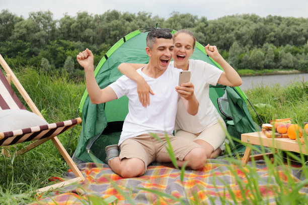 兴奋快乐的男女穿着休闲服坐在河边的帐篷里，用手机，握紧拳头，欢呼，玩耍，阅读好消息，下注和获胜。图片下载