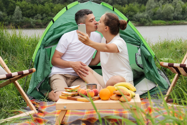 大自然中的快乐时光。在河边露营。浪漫的男女穿着休闲的衣服坐在河边的帐篷里，接吻和自拍。图片下载