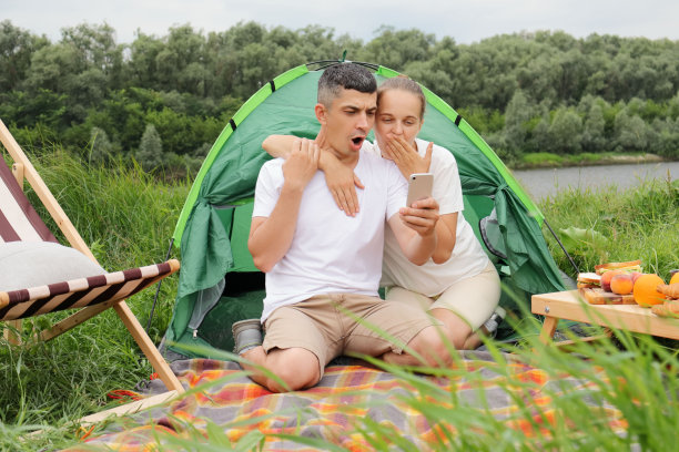 震惊不已的女子和身穿便服的男子坐在河边的帐篷旁，夫妻俩一边唱着手机，一边用惊讶的表情看着显示屏。图片下载