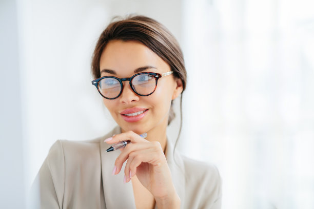成功的温柔年轻女子的特写镜头，梳着头发，把手放在下巴附近，拿着笔，专注于监视器，准备业务报告，修指甲，穿着优雅，在室内工作。图片下载