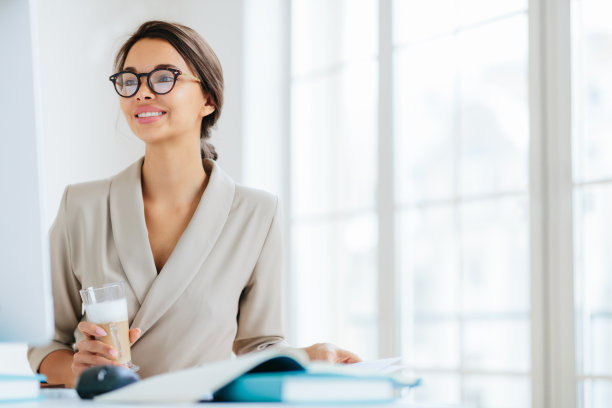 积极的女性在办公室工作，坐在办公桌前，专注于显示器，喝着新鲜的奶昔，微笑着享受工作，对着办公室内部摆姿势思考商务活动的组织图片下载